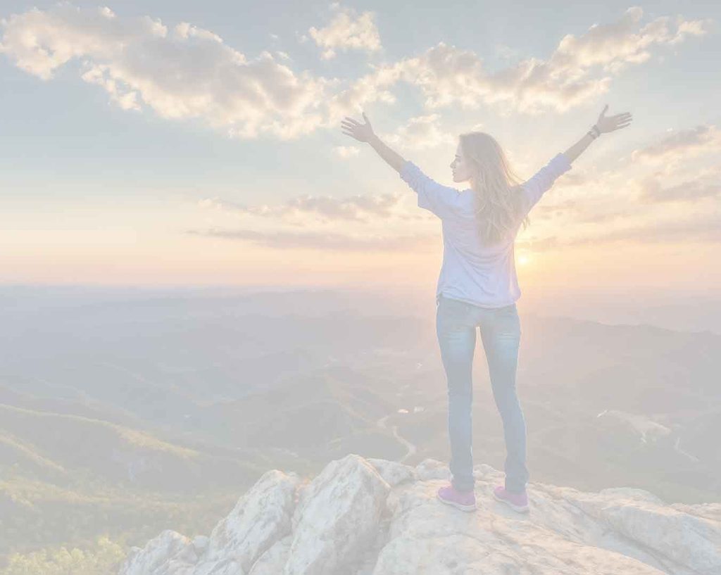 Photo of a woman standing at the top of the mountain, arms stretched toward the sunrise.