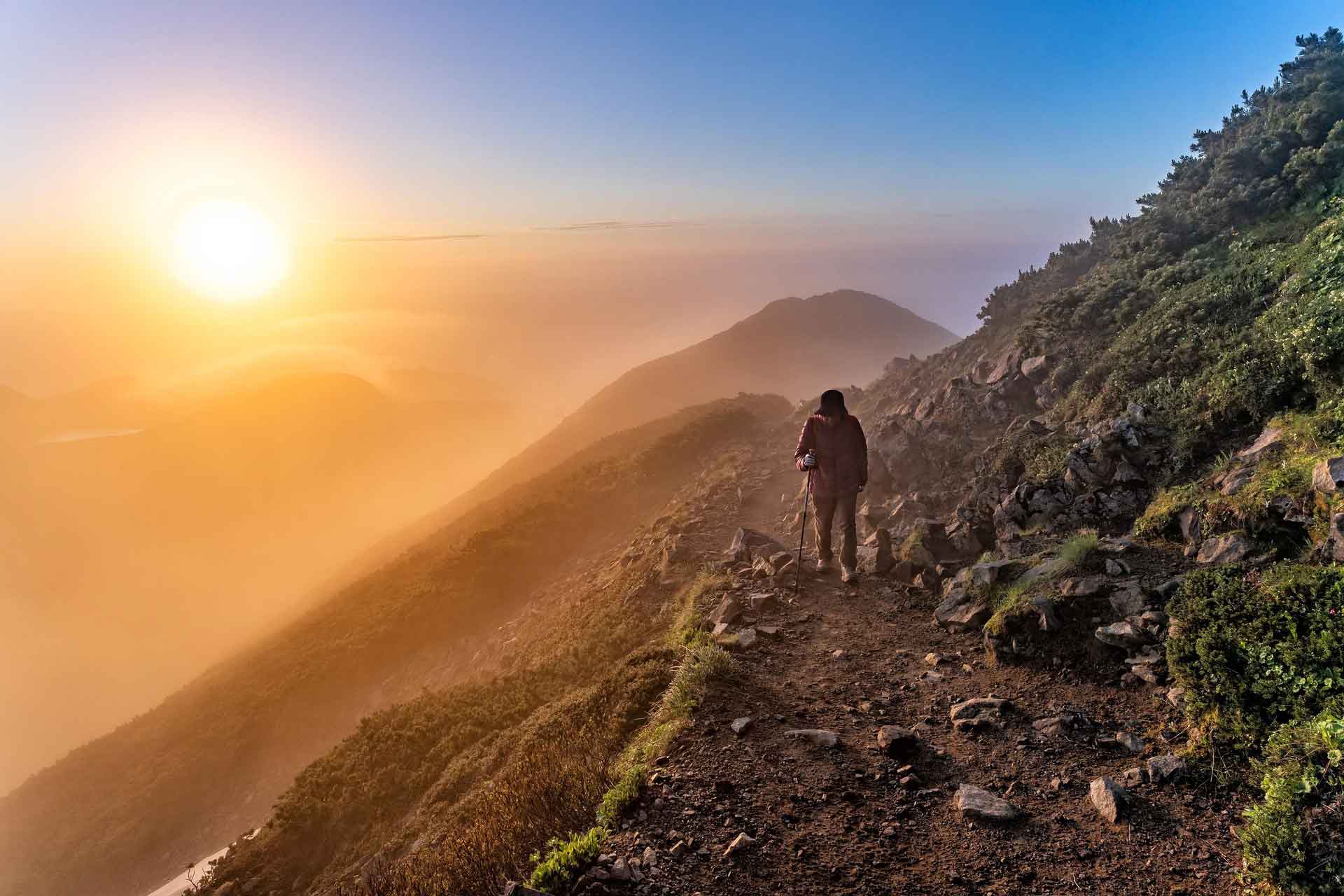 Photo of an individual hiking a steep mountainside toward the sun.