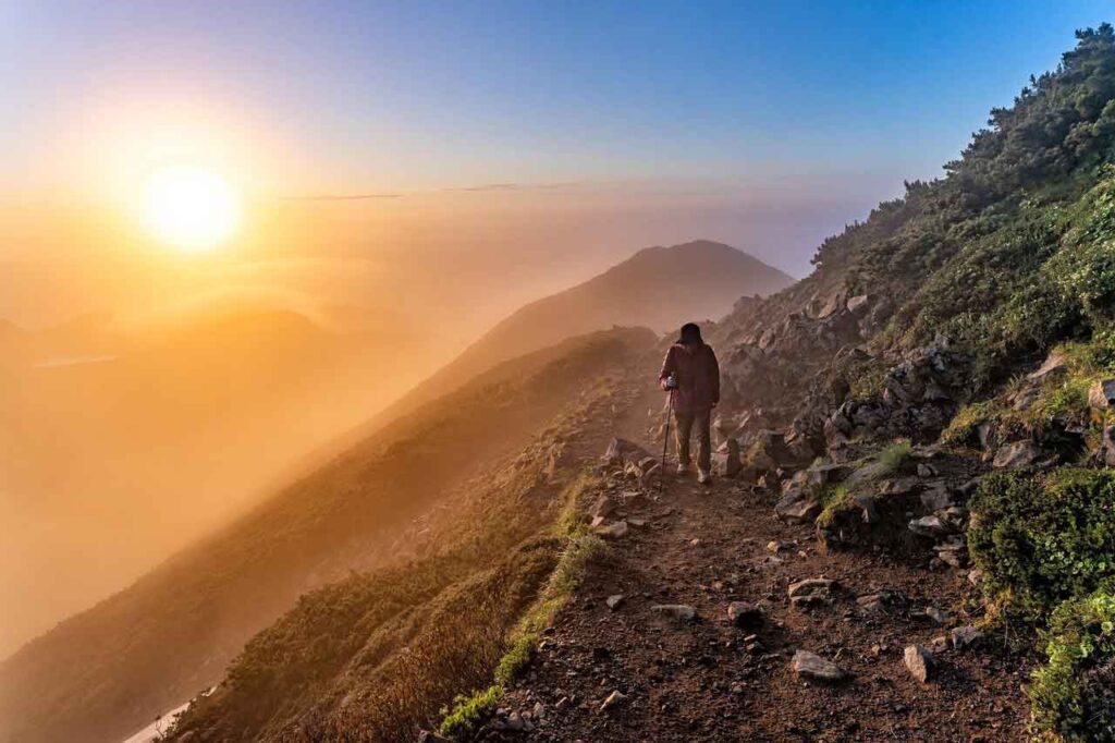 Photo of a person hiking a steep mountainside toward the sun.
