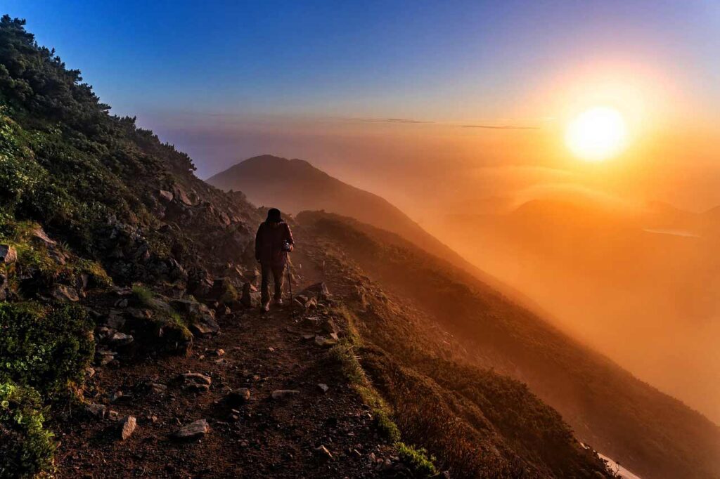 Photo of a person hiking a steep mountainside toward the sun.